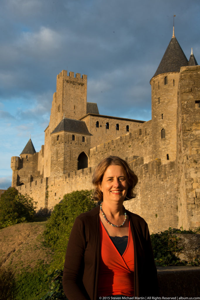 Waist-up portrait of a visitor with Carcassonne’s crenellated square tower and round roofed tower behind her.