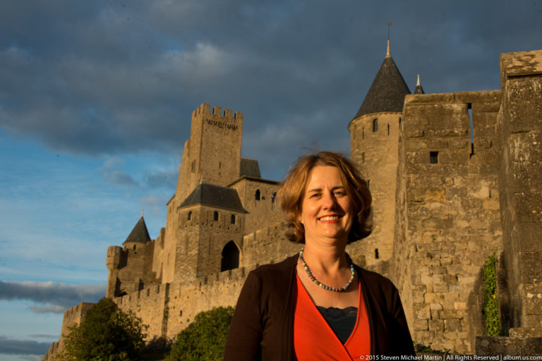 Smiling visitor stands before Carcassonne’s massive stone walls, square tower, round roofed tower, and arched opening.