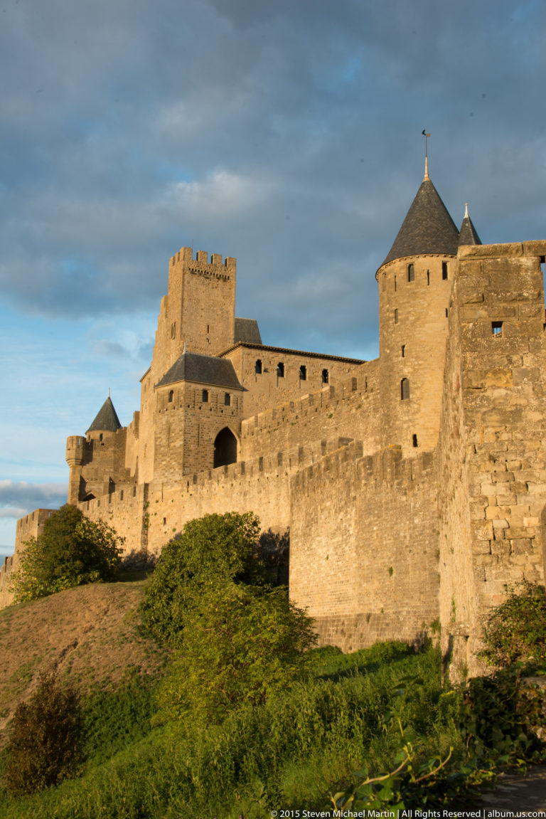 Sunlit Carcassonne walls and towers rise above dense vegetation beneath dark storm clouds.