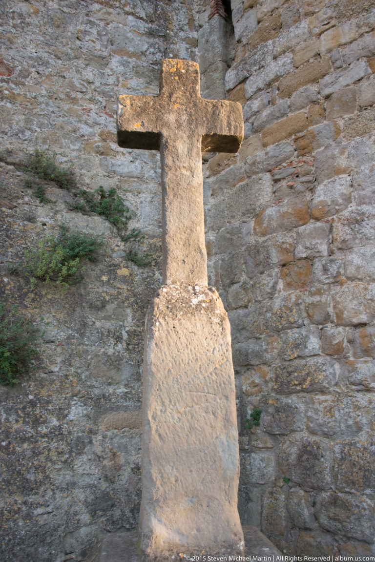 Pale stone cross-shaped marker stands against a rough masonry wall, lit by warm late-day light.