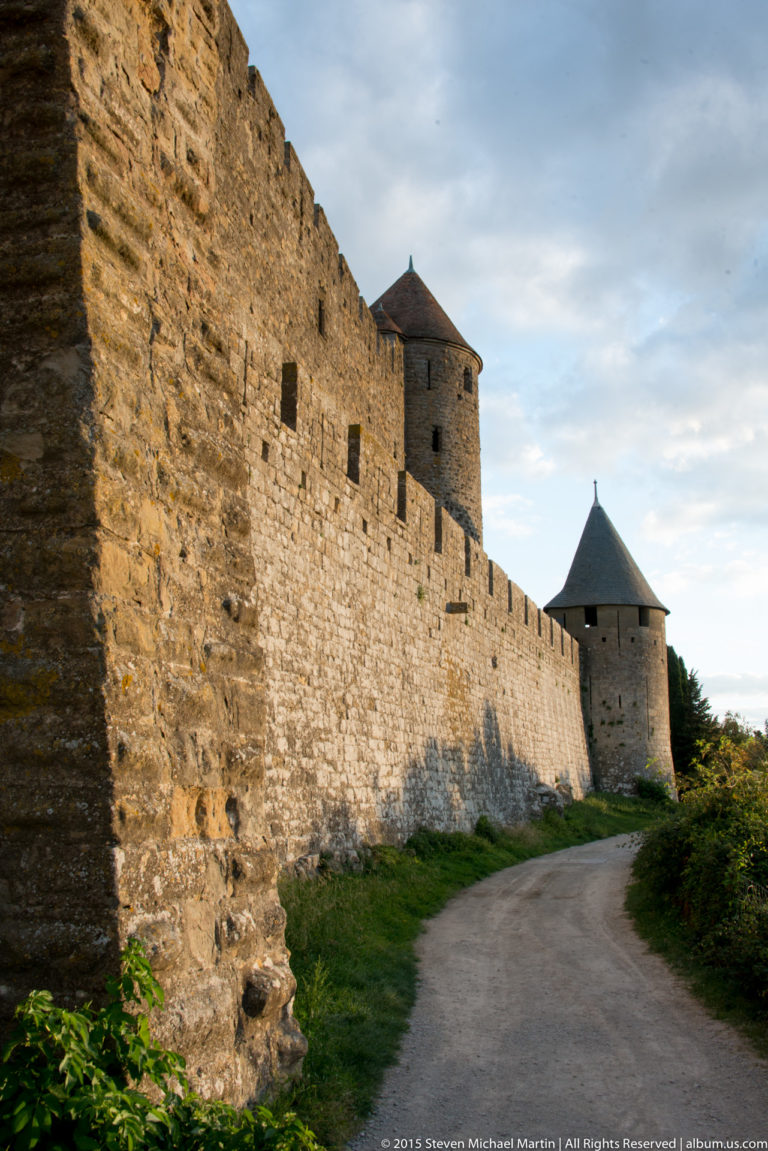 Long Carcassonne stone rampart beside a curving path, with round conical-roofed towers receding into the distance.