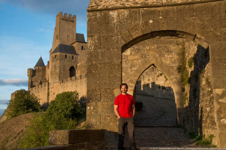 Visitor in red stands before nested stone arches, with massive Carcassonne walls and towers rising beside the passage.