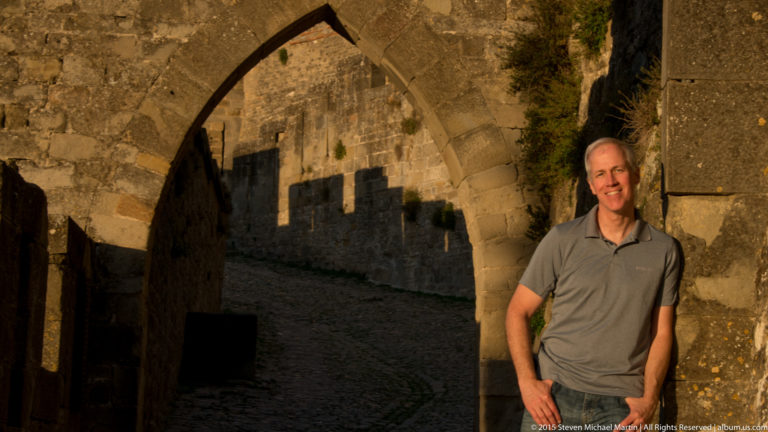 Visitor stands beside a pointed stone arch leading into a shadowed Carcassonne passage, lit by warm late-day sun.