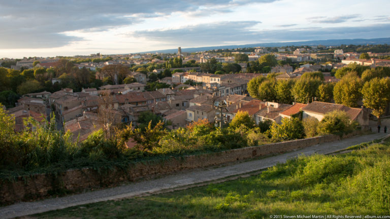 Elevated view over Carcassonne’s lower town, with tiled roofs, trees, distant fields, and hills beneath a pale evening sky.