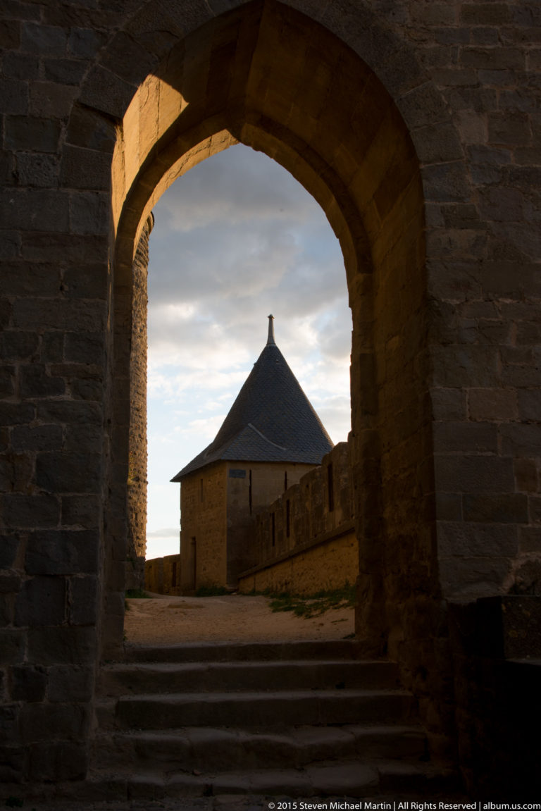 Small roofed stone structure seen through a deep archway, with steps leading upward beneath an evening sky.
