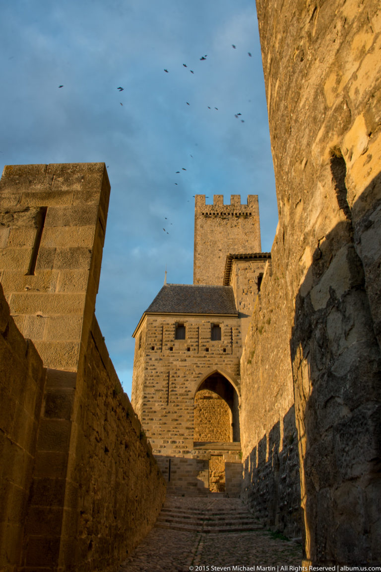 Stone steps and passage rise between high defensive walls toward an arched gateway and square tower at Carcassonne.