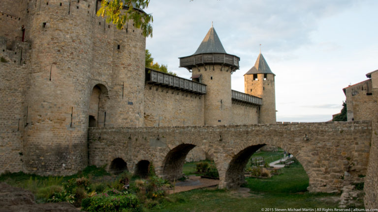 Stone bridge with several arches crosses between Carcassonne walls, with round and square roofed towers rising behind.