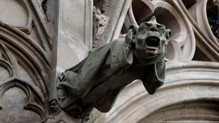 Dark stone gargoyle with open mouth and folded limbs projects from carved Gothic masonry and tracery.