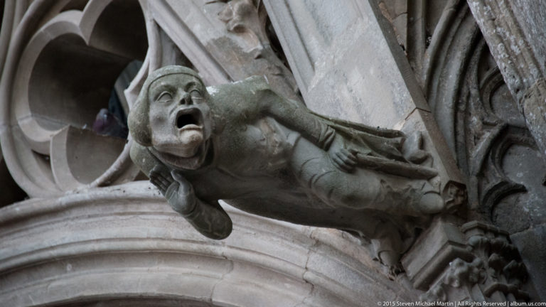 Open-mouthed crouching stone gargoyle projects from Gothic tracery and carved masonry on a church exterior.