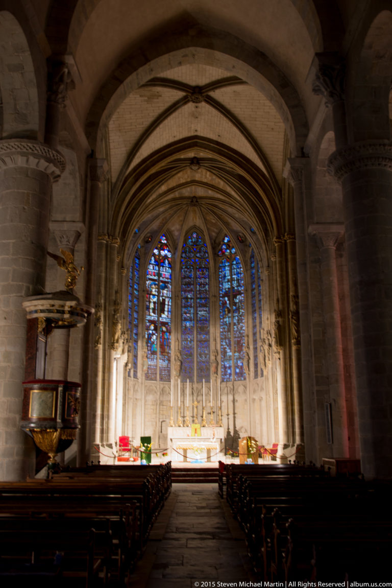 Gothic church choir with tall stained-glass windows, pointed arches, ribbed vaults, altar, and rows of dark seating.