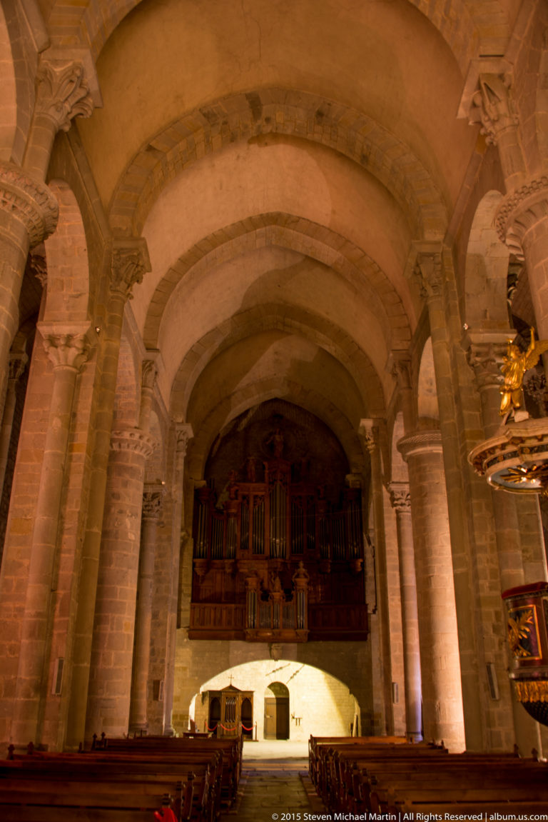 Stone church interior with heavy arches, ribbed vaulting, clustered columns, and a wooden organ or gallery at the far end.