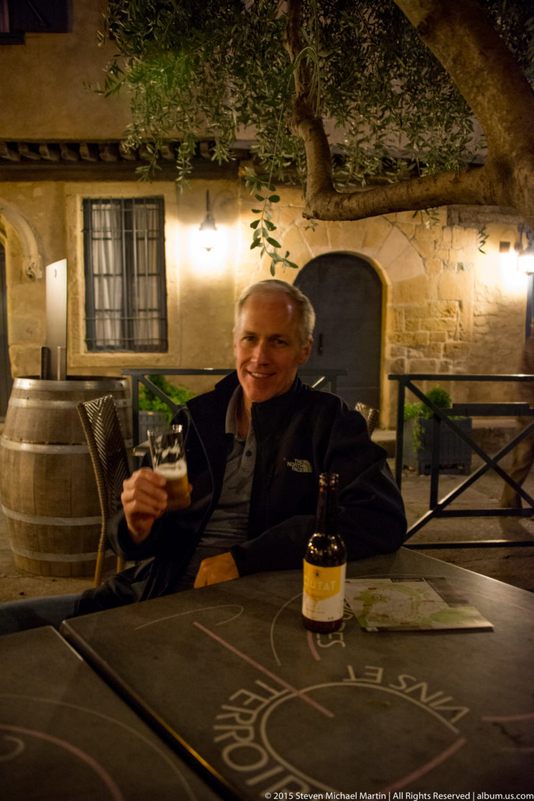 Smiling visitor holding a drink at an outdoor table with a bottle, barrel, stone wall, and tree behind him.