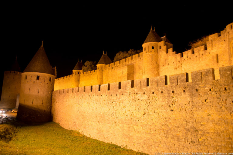 Long illuminated stone rampart leads to a round conical-roofed tower at Carcassonne under a black night sky.