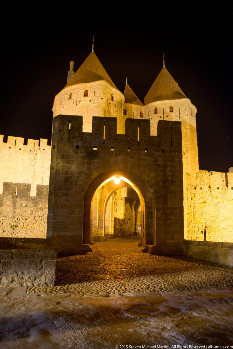 Cobblestone road enters a broad arched gateway beneath illuminated round towers and battlemented walls at Carcassonne.