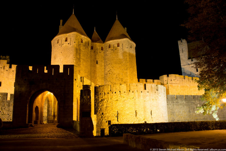 Illuminated arched gateway and paired round towers at Carcassonne, with adjoining walls and another tower visible at night.