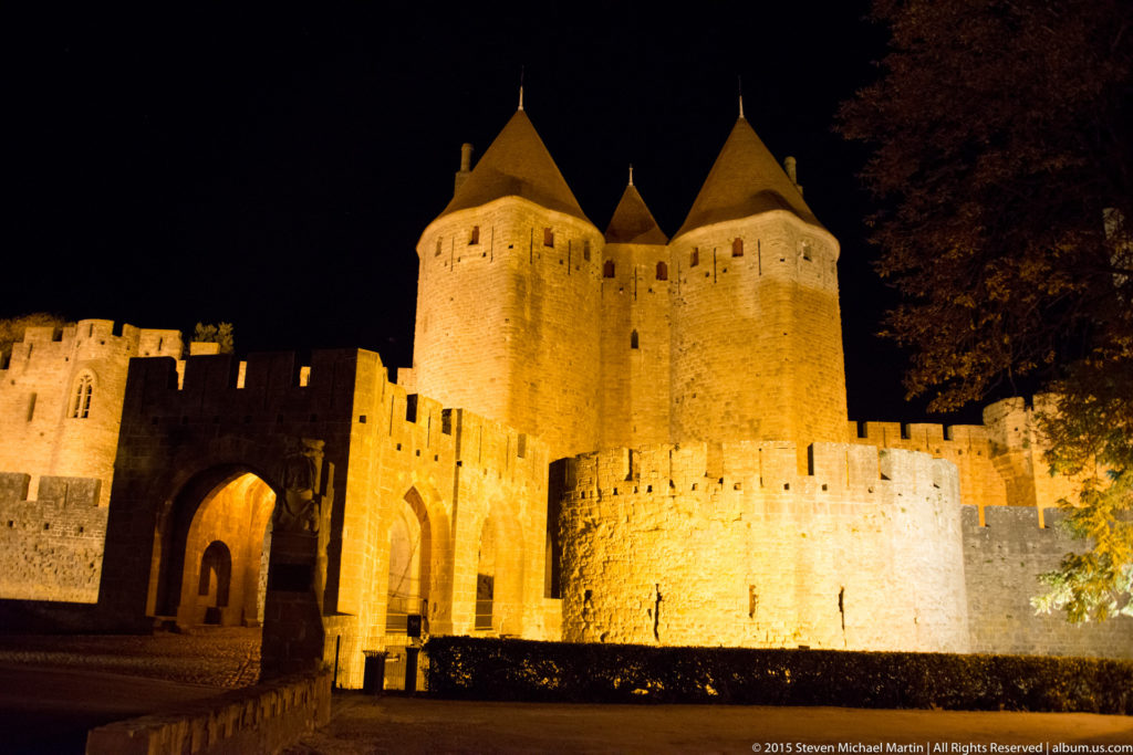 Floodlit arched gateway beneath twin round conical-roofed towers at Carcassonne, surrounded by crenellated stone walls at night.