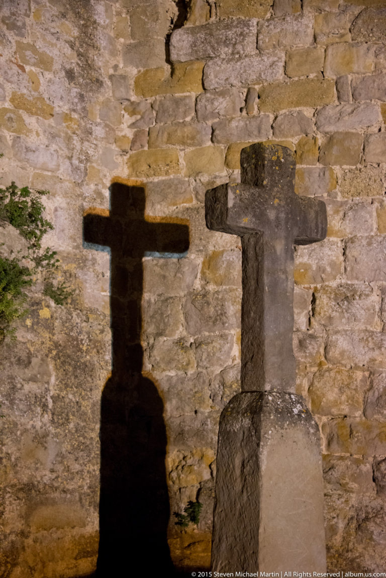 Stone cross-shaped marker beside a rough wall, casting a dark cross shadow across illuminated masonry at night.