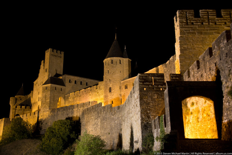Floodlit Carcassonne walls lead toward square and round towers at night, with an arched opening in the foreground.