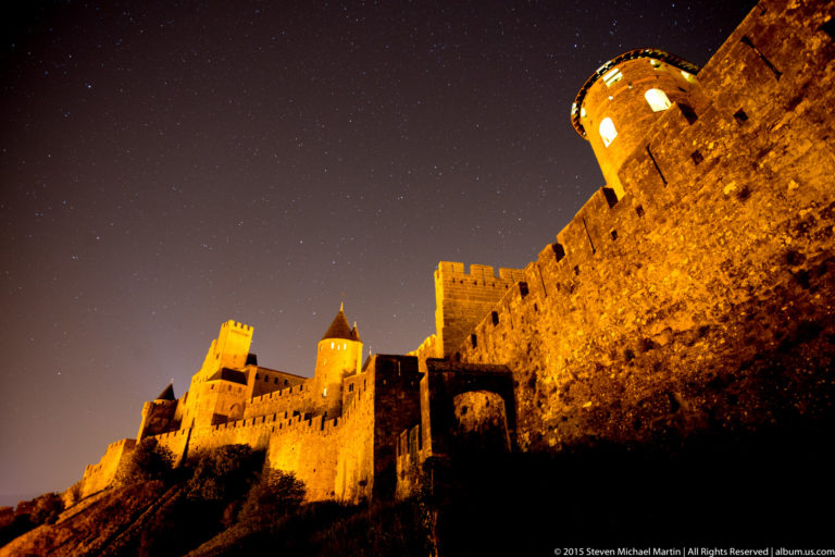 Low-angle night view of Carcassonne’s floodlit stone ramparts rising diagonally toward a round tower under a dark sky.