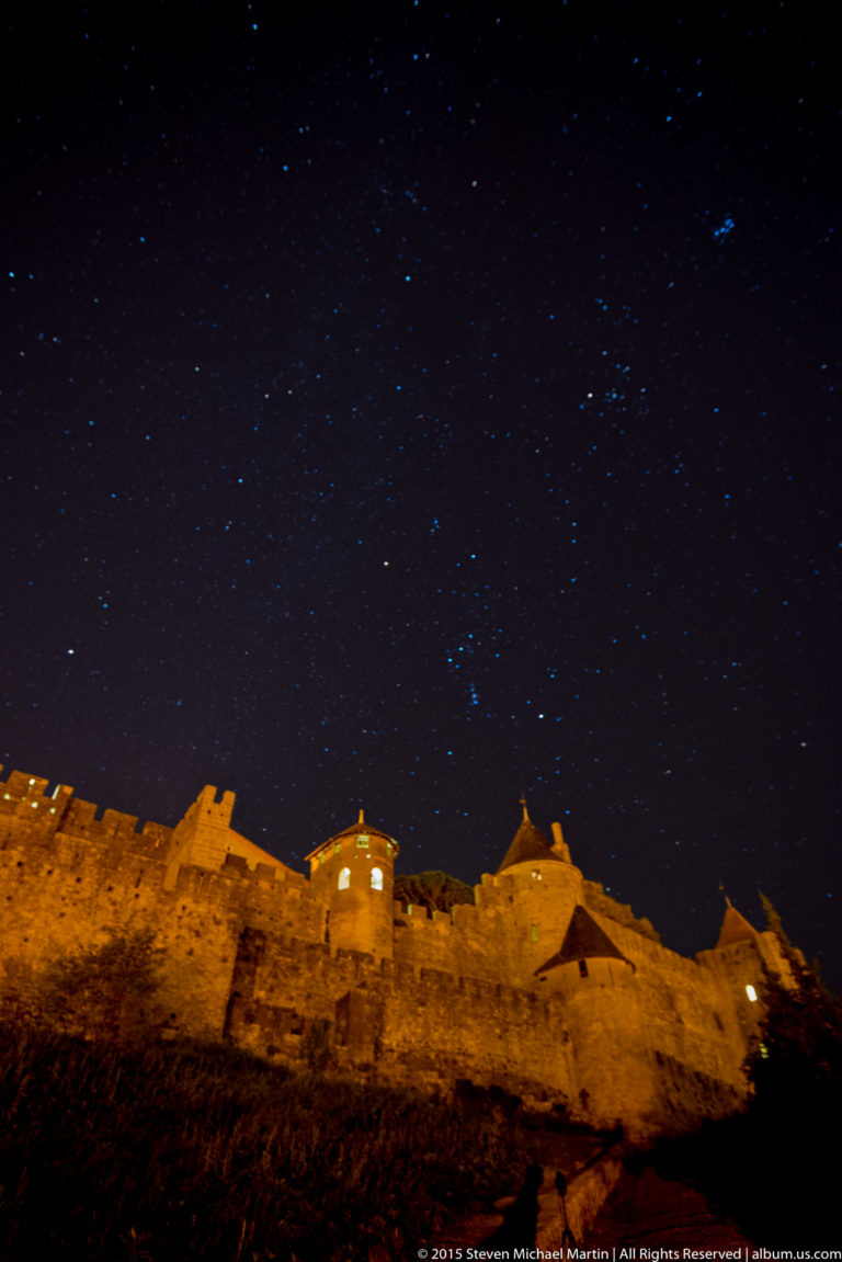 Warmly illuminated Carcassonne walls and towers form a low skyline beneath a dark sky filled with visible stars.