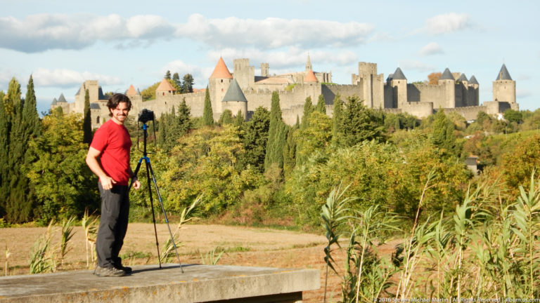 Visitor in a red shirt stands beside a tripod with Carcassonne’s towers and stone ramparts across the landscape behind him.