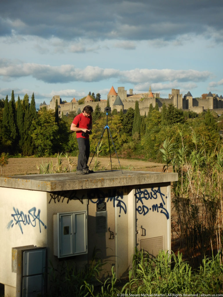 Visitor bends over a tripod on a concrete platform, with the fortified skyline of Carcassonne visible beyond trees and fields.