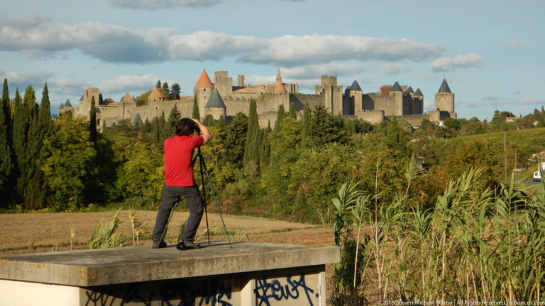 Photographer at a tripod on a concrete platform, facing the distant stone walls and towers of Carcassonne.