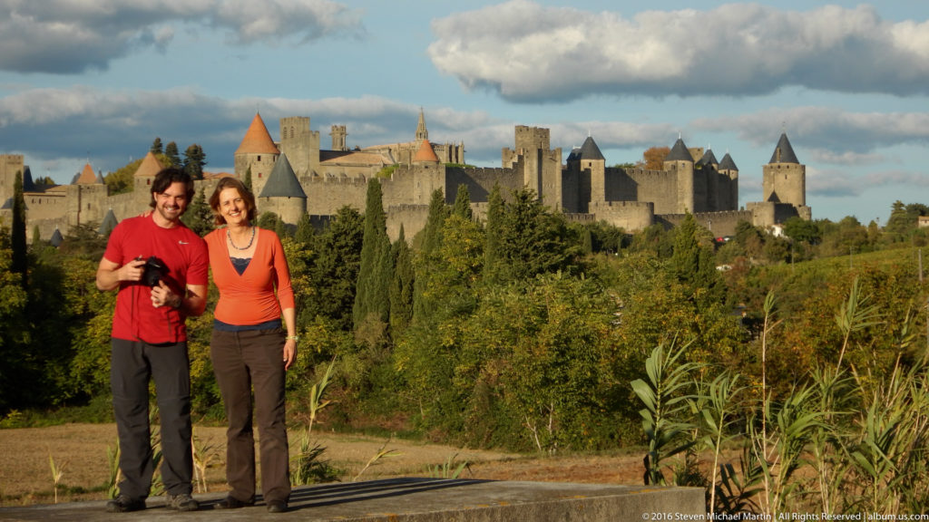 Two visitors stand at an overlook with Carcassonne’s towers, walls, and conical roofs visible across the landscape.