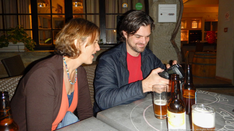 Two visitors sit at an outdoor café table with a camera, beer bottles, and glasses under warm evening light.