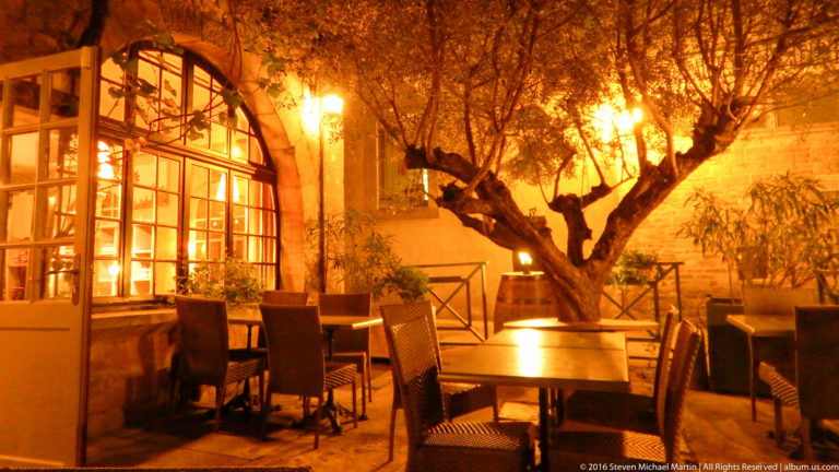 Empty outdoor restaurant tables and chairs beneath a large tree, illuminated by warm amber light at night in Carcassonne.