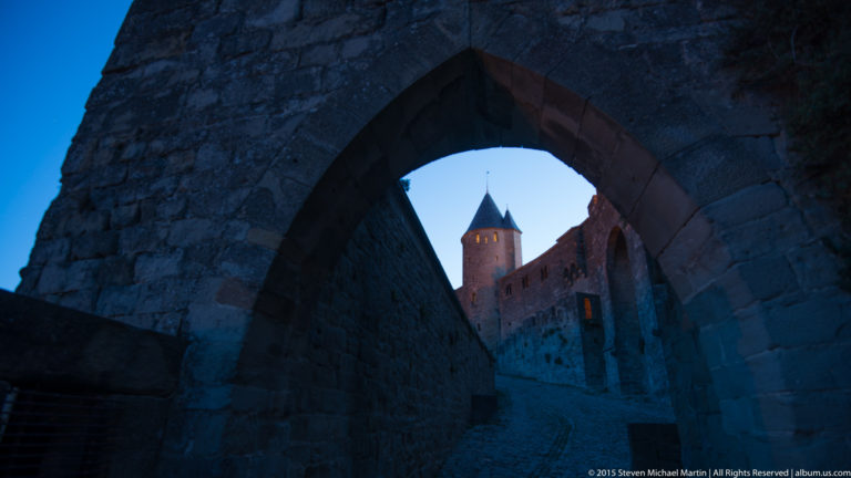 Round fortification tower at Carcassonne seen through a dark pointed stone arch in cool blue twilight.