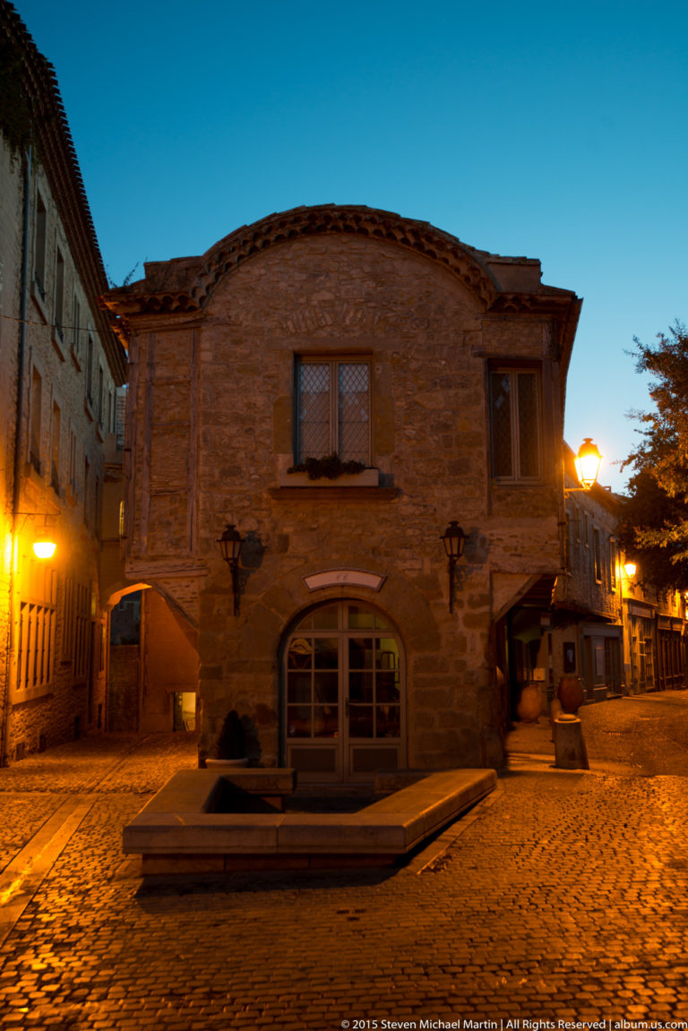 Stone building with shuttered windows faces a quiet paved square under warm lamps and a deepening blue evening sky.