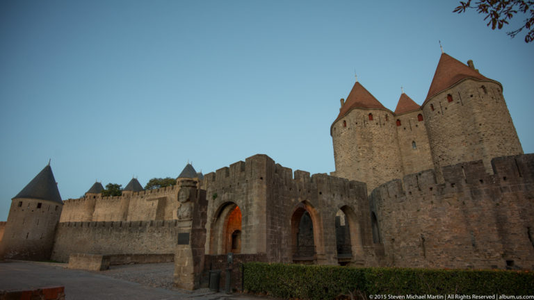 Fortified Carcassonne entrance with arched openings, twin tall towers, adjoining ramparts, and additional towers extending left.