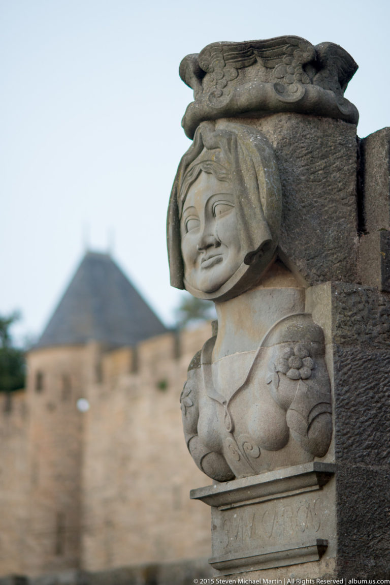 Weathered carved stone corbel with a smiling human figure, seen close-up against softly blurred masonry at Carcassonne.