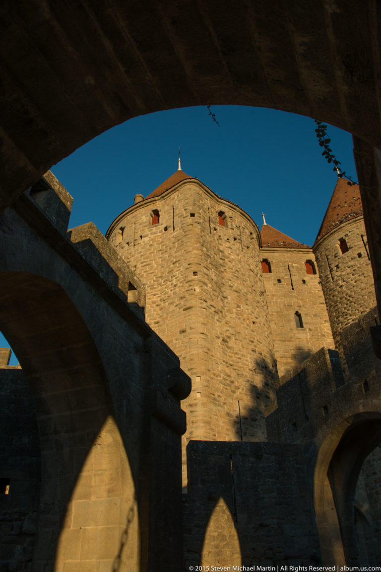 Sunlit stone wall and round fortification structure at Carcassonne seen through a deep shadowed archway.