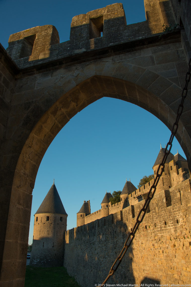 Round tower and stone ramparts at Carcassonne framed through a large masonry arch crossed by a heavy chain.