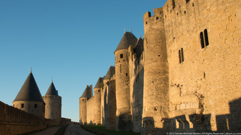 Massive sunlit stone rampart and conical-roofed towers at Carcassonne, seen from a shadowed path beside the wall.