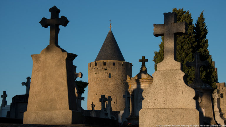 Stone cemetery crosses and grave monuments stand before a round conical-roofed fortification tower in Carcassonne.