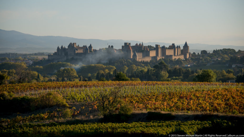 Distant Carcassonne skyline beyond vineyard rows and trees, with haze and a small plume of smoke near the walls.
