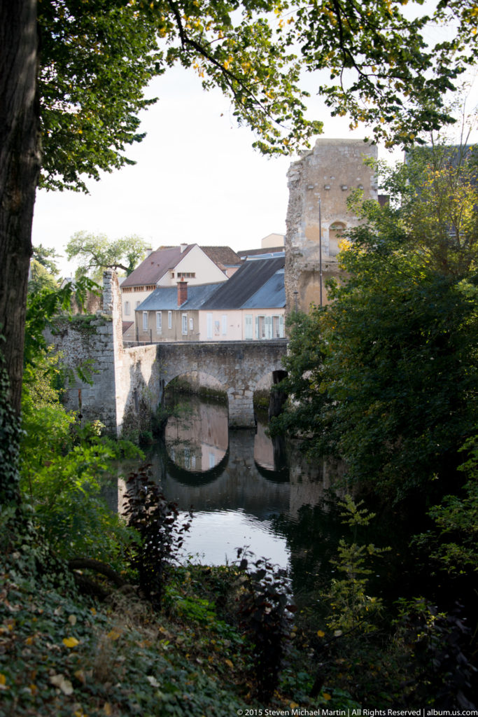 Stone bridge over the Eure River beside historic buildings and masonry walls, framed by leafy trees in Chartres.