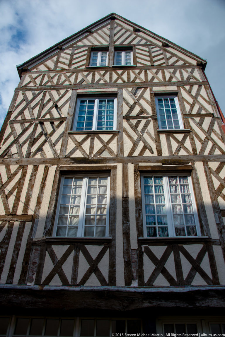 Upward view of a steep-gabled half-timbered house with dark wooden framing, pale infill, and shuttered windows in Chartres.