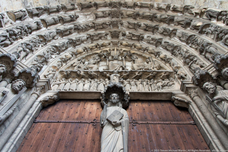 Carved Gothic portal at Chartres Cathedral with a central standing figure, wooden doors, tympanum, and crowded sculpted archivolts.
