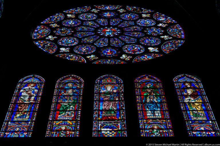 Large stained-glass rose window above five tall lancet windows glowing blue and red inside Chartres Cathedral.