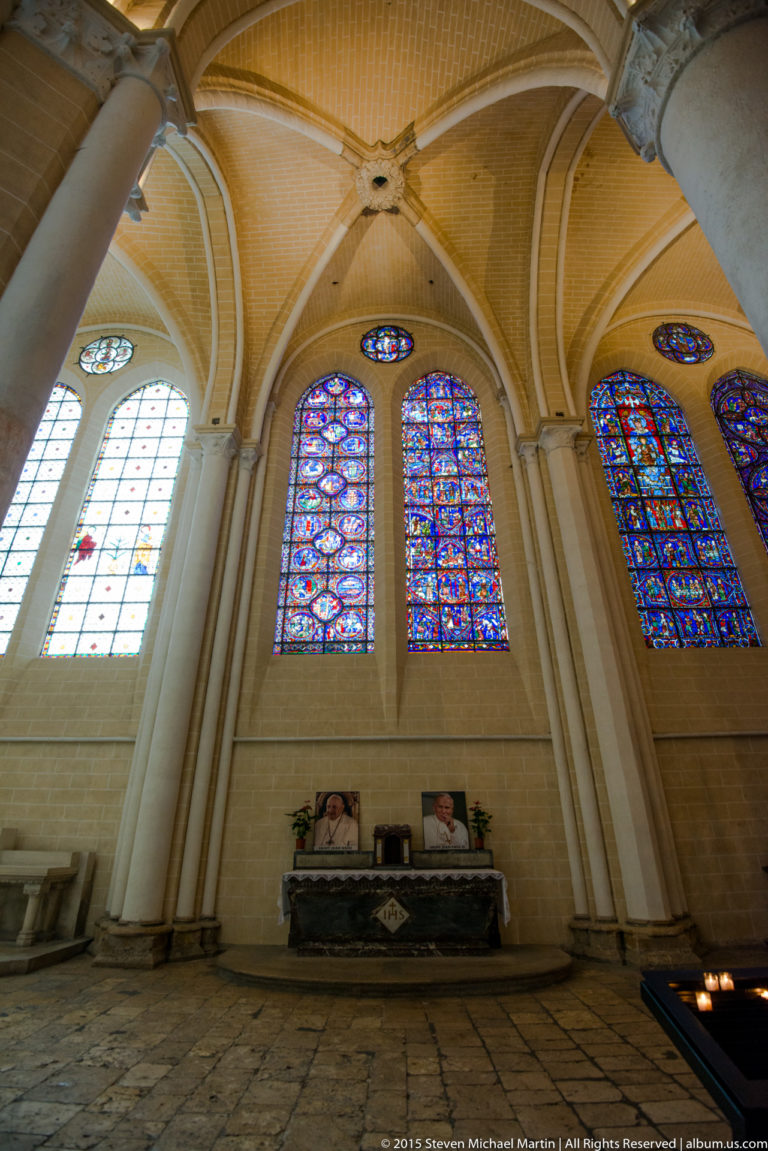 Wide interior view of three stained-glass window groups set high in the pale stone walls of Chartres Cathedral.