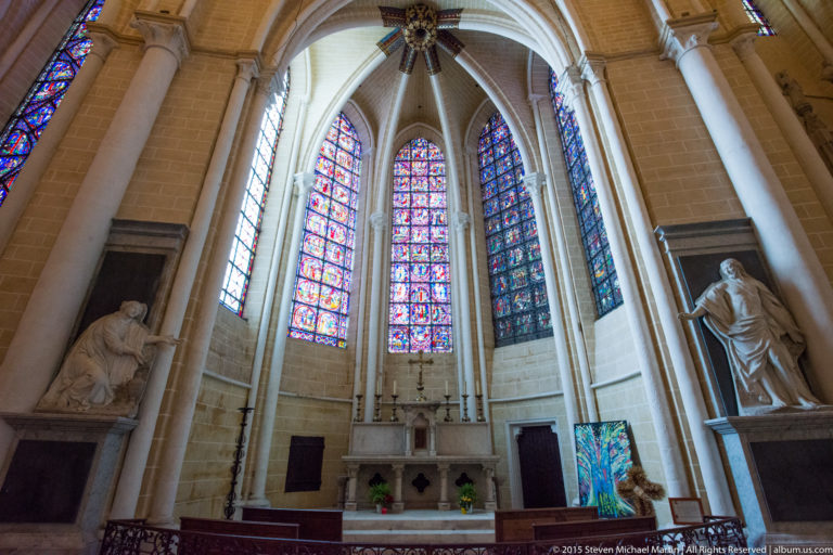 Side chapel with altar, tall stained-glass lancets, pale stone columns, and large statues in Chartres Cathedral.