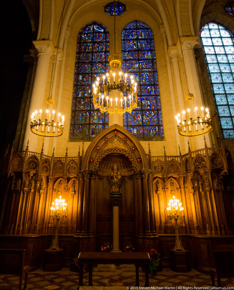Carved wooden choir stalls lit by chandeliers beneath stained-glass windows inside Chartres Cathedral.