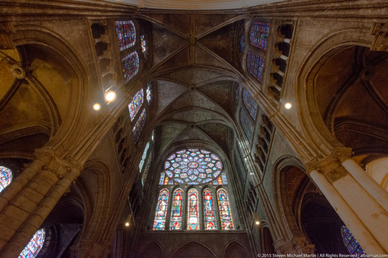 Stained-glass rose and lancet windows framed by massive arches and ribbed vaults inside Chartres Cathedral.
