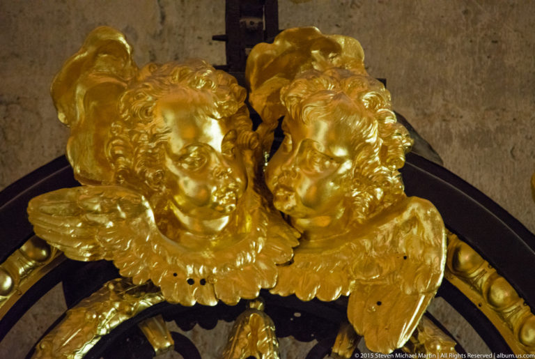 Two gilded cherub heads with small wings facing one another above a dark curved church furnishing.