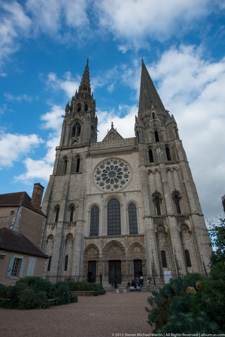Broad frontal view of Chartres Cathedral with two contrasting spires, central rose window, three portals, and pale stone façade.