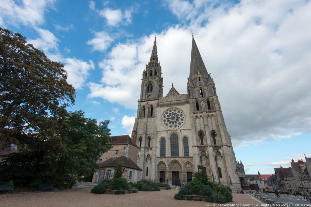 Chartres Cathedral viewed across its forecourt, with twin spires, rose window, neighboring buildings, trees, and a cloudy blue sky.
